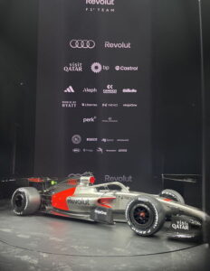 Ein silber-rotes Formel 1 Rennfahrzeug von Audi bei der Präsentation in Berlin vor einer Plakatwand mit Sponsorenlogos.